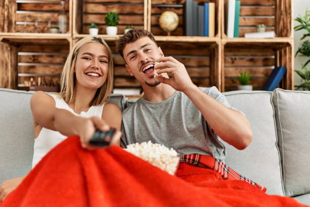 joven pareja caucásica sonriendo feliz viendo películas comiendo palomitas de maíz en casa. - apartment television family couple fotografías e imágenes de stock