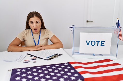 Young brunette woman at political election sitting by ballot scared and amazed with open mouth for surprise, disbelief face