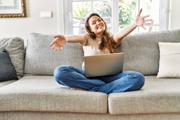 istock Beautiful young brunette woman sitting on the sofa using computer laptop at home looking at the camera smiling with open arms for hug. cheerful expression embracing happiness. 1337224536