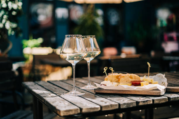 una mesa de madera en un restaurante con un plato de queso, uva, pan de miel y vino blanco. copas de vino y queso servidos para una fiesta en un bar borroso o un restaurante en el patio en un soleado día de verano - wine bottle food wine restaurant fotografías e imágenes de stock