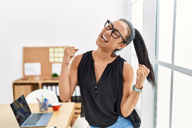 junge hispanische geschäftsfrau, die im büro arbeitet, sehr glücklich und aufgeregt, die gewinnergeste mit erhobenen armen macht, lächelt und schreit nach erfolg. feierkonzept. - women celebration excitement ecstatic stock-fotos und bilder