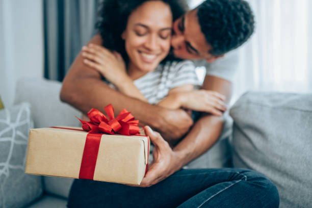 Cheerful young woman receiving a gift from her boyfriend. Shot of a loving husband giving his wife a gift. Boyfriend surprise his beautiful girlfriend with present while she is sitting on the sofa in the living room at home. Focus is on the gift. anniversary gifts stock pictures, royalty-free photos & images