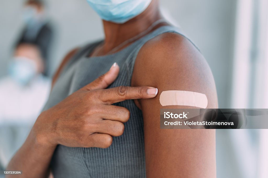 Businesswoman showing her arm after getting vaccinated in the office. Cropped shot of an attractive african-american businesswoman pointing at her arm with a bandage after receiving COVID-19 vaccine in the office with her colleagues in background. Young businesswoman showing her shoulder after getting coronavirus vaccine. Vaccination Stock Photo