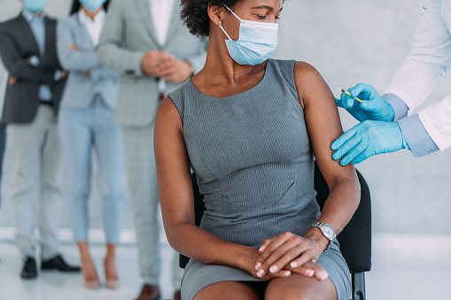Shot of an attractive african-american businesswoman getting vaccinated in the office with her colleagues in background. Male doctor giving a vaccine of young woman. Shot of general practitioner giving injection to young woman at vaccination center. Female patient getting vaccinated during Covid-19 pandemic. Coronavirus vaccination concept.