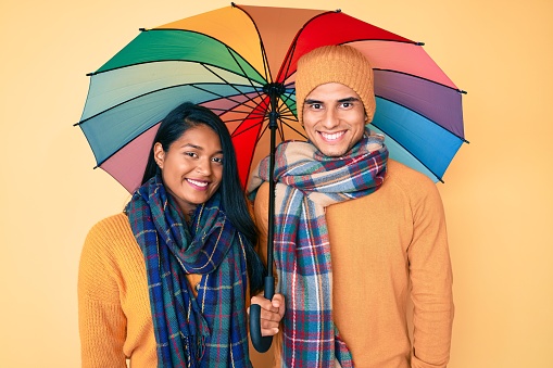 Beautiful latin young couple under colorful umbrella looking positive and happy standing and smiling with a confident smile showing teeth