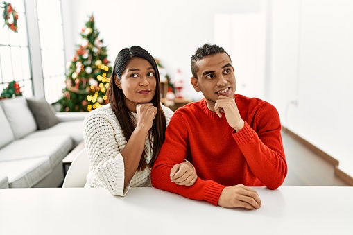 Young latin couple sitting on the table by christmas tree with hand on chin thinking about question, pensive expression. smiling and thoughtful face. doubt concept.