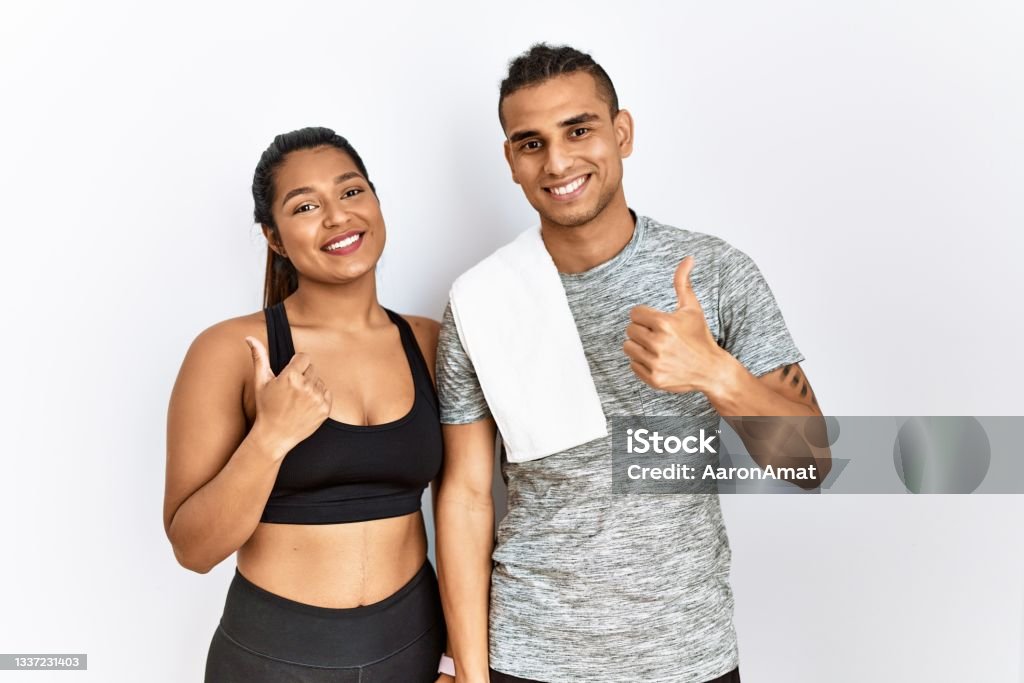 Young latin couple wearing sportswear standing over isolated background doing happy thumbs up gesture with hand. approving expression looking at the camera showing success. Exercising Stock Photo