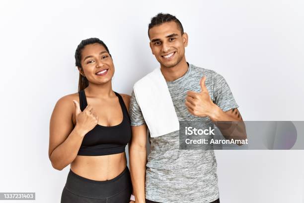 Young Latin Couple Wearing Sportswear Standing Over Isolated Background Doing Happy Thumbs Up Gesture With Hand Approving Expression Looking At The Camera Showing Success Stock Photo - Download Image Now