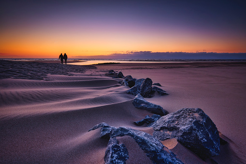 A sandy beach during sunset. There is a path of stones going into the sea and 2 people are walking over them. The sky is beautifully colored by the setting sun.