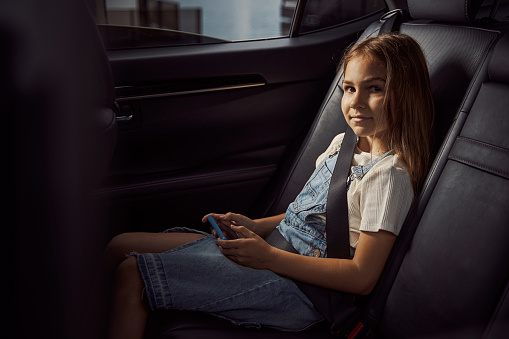 Cheerful pretty girl holding a smartphone and smiling while sitting in a car with a safety belt fastened