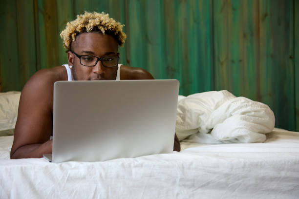 African-American man is a wide-awake man in a white T-shirt and glasses working on a modern laptop in bed. Always online.