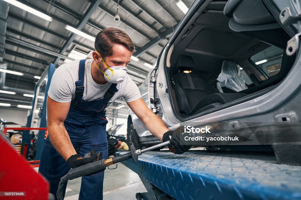 Mechanic removing dents from rear part of car Repair service man in respirator using slide hammer for peening of car rear bumper Dented Stock Photo