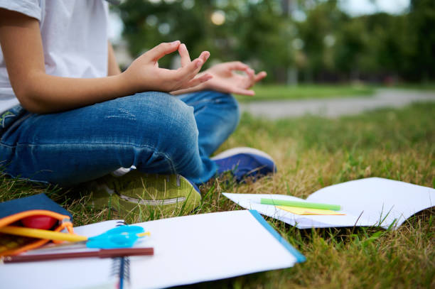 Cropped view of school child boy sitting in lotus position on green grass of city park and meditating . Workbooks school supplies lying on the grass. Concentration, recreation, mindfulness concepts stock photo