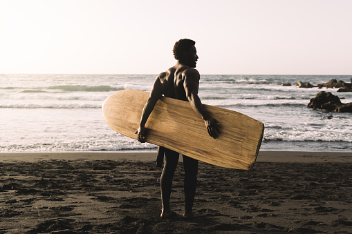 Black surfer man holding vintage surf board on the beach at sunset - Focus on back