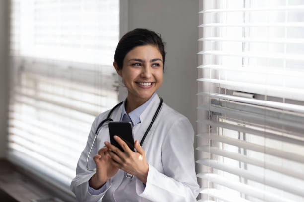smiling dreamy young indian doctor holding cellphone in hands. - médico geral imagens e fotografias de stock
