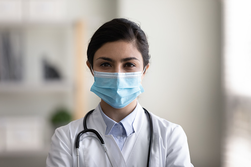 Head shot portrait of confident female indian ethnicity skilled infectious disease specialist in medical uniform and disposable protective facial mask looking in camera, posing in clinic office.