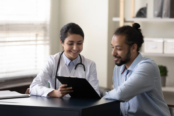 Happy young indian doctor discussing treatment with african patient. Happy young indian female gp doctor showing test result on clipboard to interested millennial african ethnicity male patient, discussing healthcare medical insurance or illness treatment at clinic. clipboard photos stock pictures, royalty-free photos & images