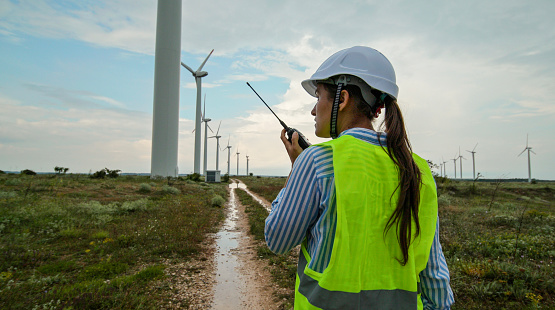 Electrical maintenance engineer working for the energy industry, supervising the condition of the power equipment in a wind turbines power station on a rainy day. Checking the data and the results of measurements with digital tablet. Woman engineer working on the field. Technology and Global Business.