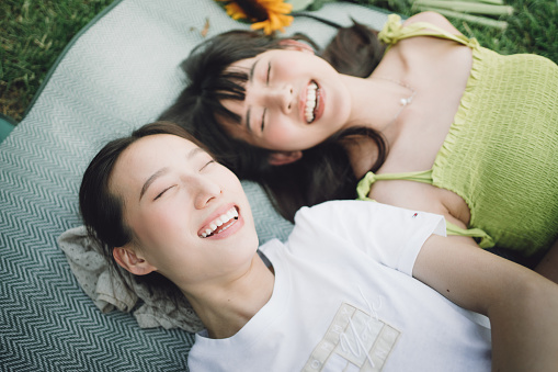 Two Asian young cheerful women lying down on the grass in the park to enjoy the sunshine in springtime.
