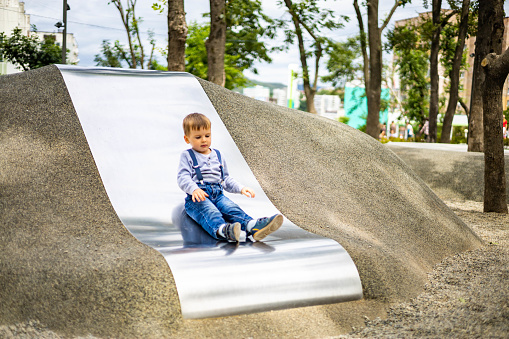 Cute baby boy riding on metallic slide at childish playground relaxing outdoor having summer vacation. Cheerful casual male baby preschooler spending time at amusement park enjoying childhood
