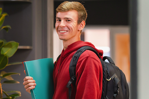 Side view photo of a happy student man in university wearing red hoodie with backpack on the back holding the color folder in his hand. High quality photo