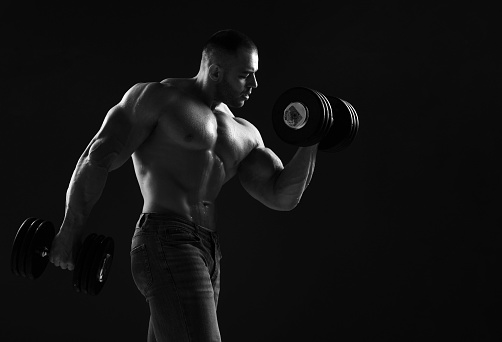 Muscular men is working out in gym, lifting alternately two big heavy dumbbells, doing exercises for biceps over black background with copy space. Young man lifting weights. Black and white