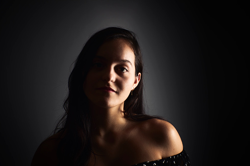 Close-up portrait of a young woman with a positive gaze looking at the camera, on a dark studio background. Happiness concept