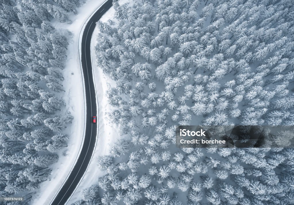 Driving In Snowcapped Forest Red car driving on black asphalt road leading through snowcapped winter forest. Aerial view. Winter Stock Photo