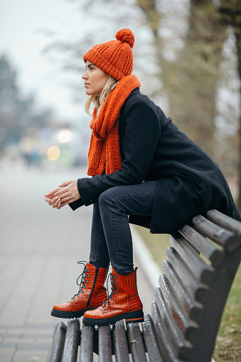 Beautiful young woman sitting on bench in city park. She wears warm clothes, shoes, scarf and cap and looks very fashionable