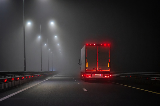 The truck is driving along the night road in the fog. Rear red lights are visible. There is street lighting.