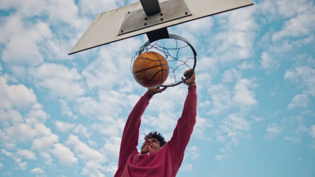 Slow motion shot of a young basketball player thrusts the ball forcefully down through the basket on the basketball court. He performs a slam dunk. Low angle shot under the basketball hoop.