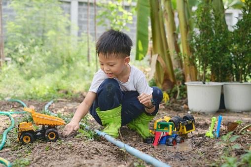 Cute Asian young schoolboy wearing rain boots playing in muddy puddles alone, Children getting dirty while digging in muddy soil with toy construction machinery in backyard home garden on nature