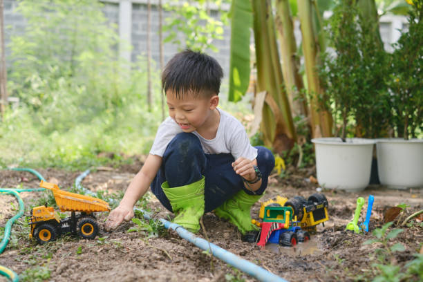 Asian boy wearing boots playing in muddy puddles, digging in mud Cute Asian young schoolboy wearing rain boots playing in muddy puddles alone, Children getting dirty while digging in muddy soil with toy construction machinery in backyard home garden on nature kids dirty shoes stock pictures, royalty-free photos & images