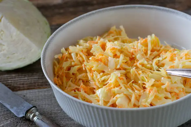 Delicious traditional, american coleslaw made with grated white cabbage, carrot, onions and a salad dressing with mayonnaise. Served in a white bowl on rustic and wooden table background.