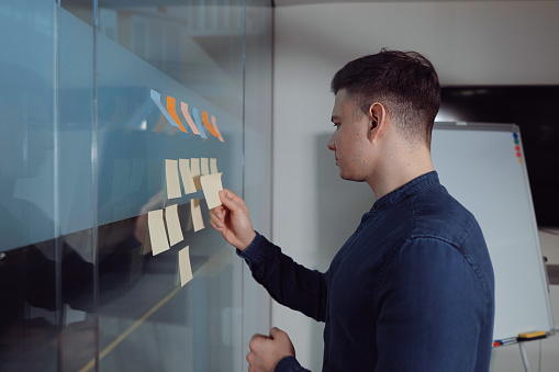 Young man wearing denim shirt checking and rearranging sticky notes on glass wall in office. Side view project manager working with Kanban board and controlling workflow. Concept of planning