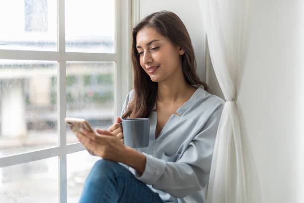 Pretty young caucasian woman sitting sofa enjoying first morning coffee on sunshine and use smartphone to check news. her enjoying pleasant memories. daily routine and relaxation time stock photo