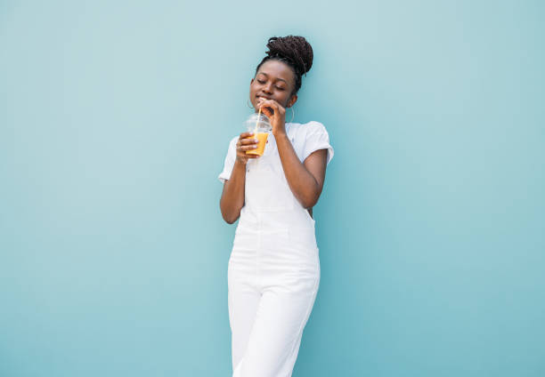 istock Young woman in white clothes drinking orange juice while standing at blue wall 1337414572