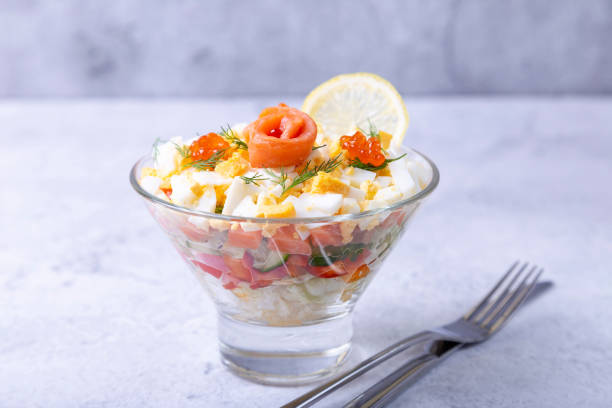 Puff salad with salted trout (salmon), caviar, egg and vegetables. A traditional dish, one serving in a glass glass on a gray background. Croup