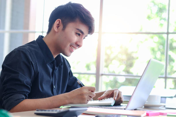 istock Asian young handsome businessman in black shirt working on office desk, taking notes and using laptop computer, writing information data and new idea on paper. 1337422919