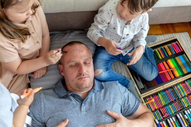 Adorable kids having fun with a sleeping father at home. Adorable kids having fun with a sleeping father at home. Kids are having fun drawing on their father's face while he his sleeping on the floor. April fools prank. Lifestyle. April Fools Pranks stock pictures, royalty-free photos & images