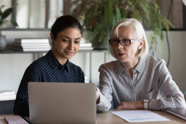 Serious mature businesswoman mentor teaching intern, using laptop