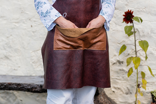 A low angle front view of an unrecognizable woman with her hands in her handmade leather apron which has been made by the owner using ethically sourced leather and sustainable products. She is stood outdoors outside of her design studio in Wales. / Female Focus Collection