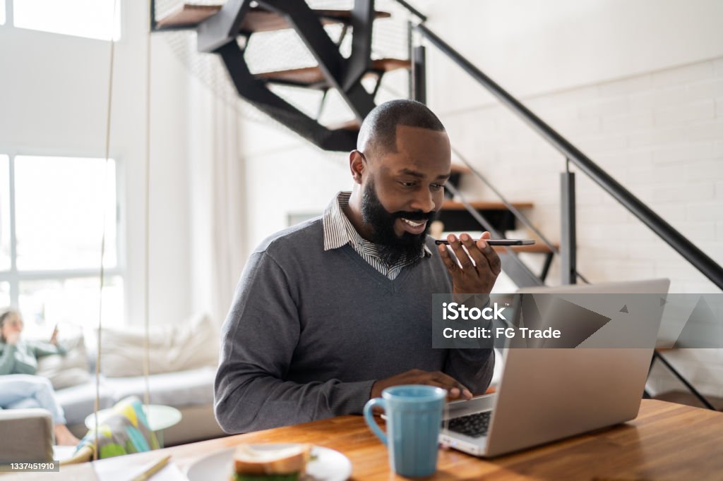 Man using laptop and sending audio message on smartphone at home People Stock Photo
