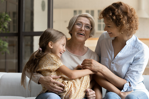Happy little girl with smiling mother and laughing mature grandmother in glasses having fun hugging sitting on couch at home, excited three generations of women, family enjoying leisure time together