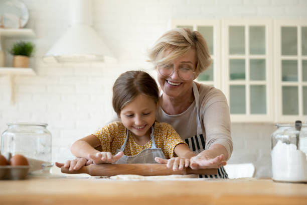 felice nonna matura che insegna adorabile nipotina a rotolare l'impasto - grandmother senior adult family domestic kitchen foto e immagini stock