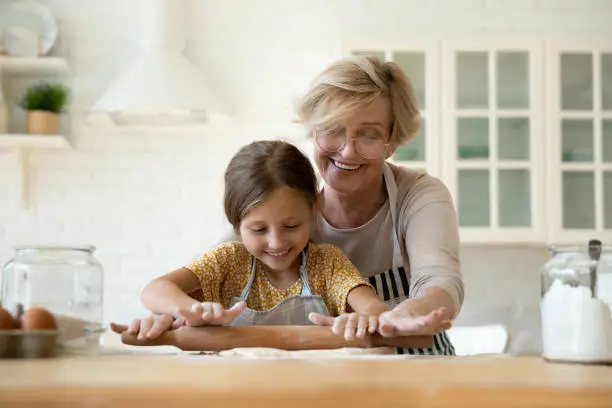 Happy mature grandmother teaching adorable little granddaughter to rolling dough, smiling senior woman in glasses with preschool girl cooking homemade cookies together, family enjoying leisure time