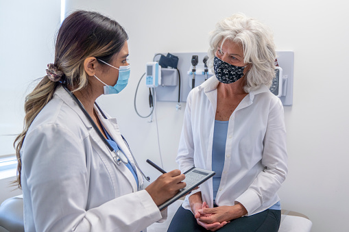 A senior woman is seated on an examination table at a doctor's office. Her female doctor is standing in front of her while holding a pen and a clipboard to write notes. They are both wearing a face mask to prevent the spread of germs. She is listening intently as her doctor is speaking to her.
