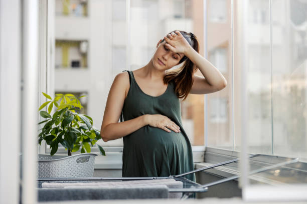 Portrait of a pregnant young woman tired of housework. The tortured woman keeps one hand on her stomach while the other wipes the sweat from her forehead, tired of obligations Balcony, day, summertime stock photo