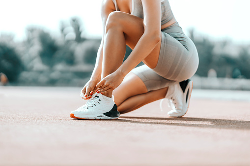 Mujer deportiva atando cordones de zapatos en entrenamiento. Una mujer con un atuendo deportivo se agacha y se ata un cordón de zapatos. Vestuario gris y zapatillas blancas en el estadio de entrenamiento al aire libre, foto cercana de un cuerpo femenino photo