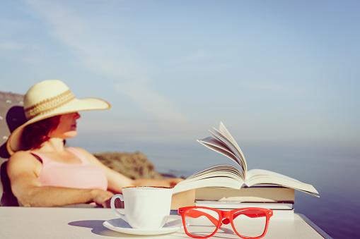 Reading on holidays. Mature tourist woman relaxing on coast, read book, enjoy sea view from cliff. Focus on coffee cup and glasses.